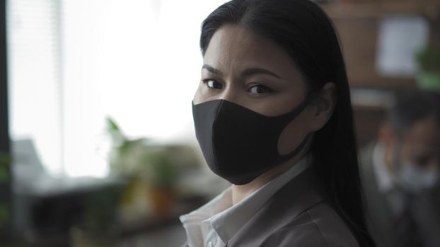 Well-Dressed Businesswoman In Black Protective Mask Looks Back At Camera, Asian Young Woman Working In Office During Quarantine, Focus On Foreground
