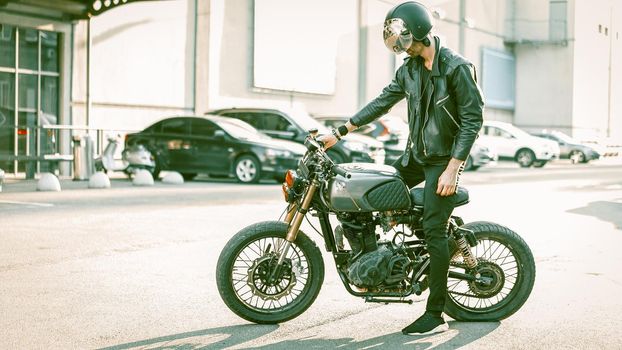 Biker In Helmet And Black Leather Wear Getting Ready To Ride, Man On Kraft Retro Style Motorbike Standing On Asphalt Road Outdoors With Shopping Center On Background, Rider About To Start Engine