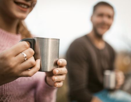 Smiling Woman Holds Metal Mug Sitting In Nature With His Man On Blurred Background, Couple Drink Hot Tea Or Coffee In Nature, Selective Focus On Females Hands Holding Steel Cup, Close Up