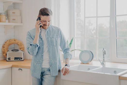 Handsome busy bearded businessman in casual comfy clothes standing in kitchen holding mobile phone, talking with client and dealing with customer issues while working remotely from home