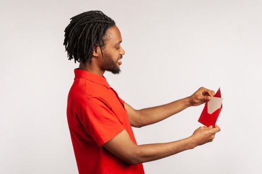 Profile of man with dreadlocks wearing red casual style T-shirt, reading letter or greeting card, holding envelope, smiling and rejoicing pleasant news. Indoor studio shot isolated on gray background.