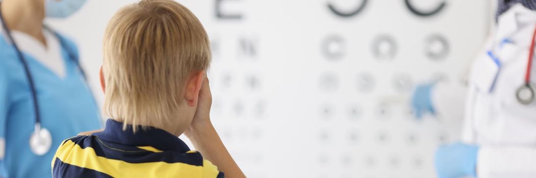 Portrait of little boy on appointment at oculist office, check sight closing eye and say out loud letter on board. Childhood, ophthalmology, health concept