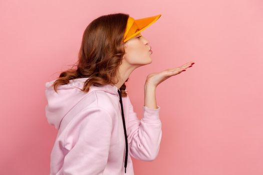 Profile of curly haired teenage girl in hoodie and yellow sun visor with plastic shield sending air kiss, flirt and affection. Indoor studio shot isolated on pink background