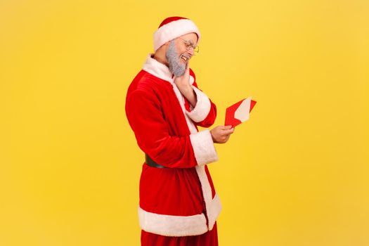 Profile portrait of elderly man with gray beard wearing santa claus costume being touched with sensitive letter, reading Christmas congratulations. Indoor studio shot isolated on yellow background.