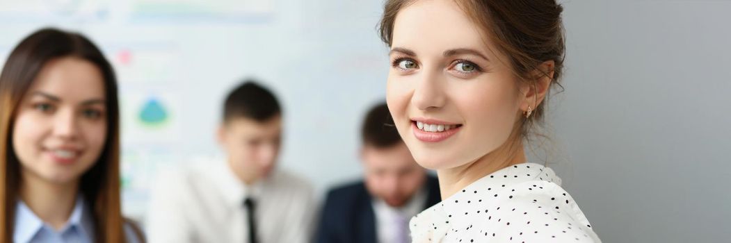 Portrait of smart woman sitting in big modern office with colleagues and smile at camera. Brunette secretary work in friendly collective. Business concept