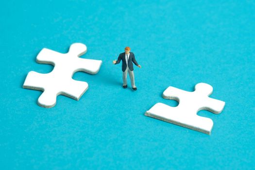 Miniature people toy figure photography. A shrugging businessman standing the middle of two puzzle jigsaw piece. Isolated on blue background. Image photo