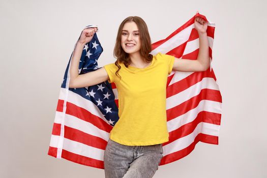 Smiling woman of young age with pleasant appearance and wavy hair, wears casual style yellow T-shirt, holding USA flag and looking at camera with smile. Indoor studio shot isolated on gray background.