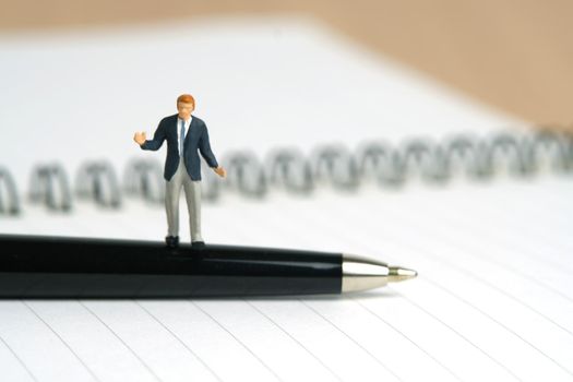 Miniature people toy figure photography. Signing agreement concept. A shrugging businessman stand above a black pen and opened document on wooden desk. Image photo