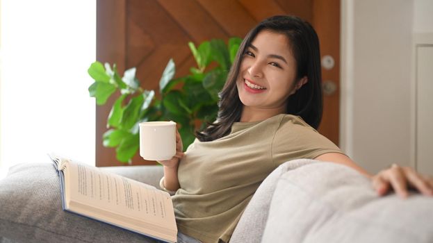 Cheerful asian woman in casual clothes holding cup of tea in her hand, relaxing on couch at home.