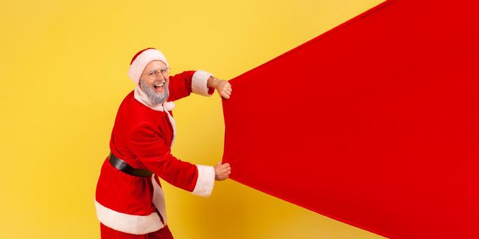 Side view portrait of elderly man with gray beard wearing santa claus costume carrying big bag with presents, copy space for advertisement. Indoor studio shot isolated on yellow background.