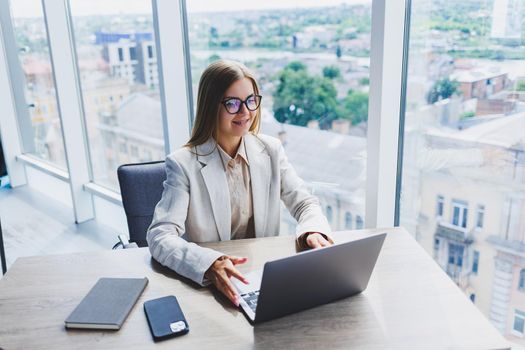 A female head of a company in glasses sits at a laptop in her office with a breathtaking view of the city. Business lady works at a wooden table.