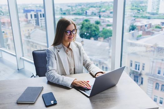 A female head of a company in glasses sits at a laptop in her office with a breathtaking view of the city. Business lady works at a wooden table.