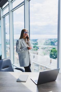 Cheerful young beautiful woman in glasses talking on a mobile phone and using a laptop with a smile while sitting at her workplace. Work in the office