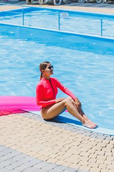 Pretty woman in a bright pink swimsuit near the pool on vacation, she is wearing sunglasses. The concept of summer holidays in the hotel by the pool. Inflatable pink mattress on the background