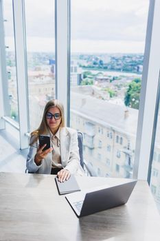 A fair-haired cheerful European woman in glasses in stylish casual clothes is sitting at a table with a laptop, doing paperwork and talking on the phone. Business lady at the workplace in the office
