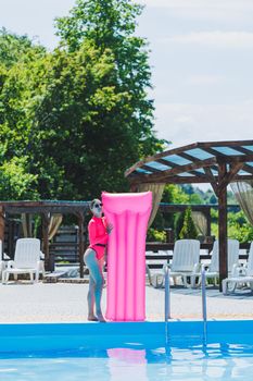 A beautiful woman in a pink swimsuit and with an inflatable mattress stands near a large pool. Summer holidays in a hotel with a swimming pool. Young girl in swimsuit and sunglasses