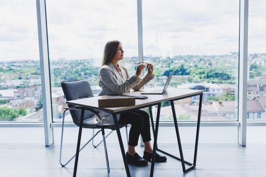 A female head of a company in glasses sits at a laptop in her office with a breathtaking view of the city. Business lady works at a wooden table.