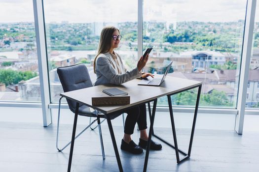 A woman manager in glasses sits at a laptop in an office with a stunning view of the city and conducts a business conversation on the phone. Business lady working at the wooden table in the office