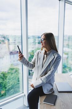 Cheerful young beautiful woman in glasses talking on a mobile phone and using a laptop with a smile while sitting at her workplace. Work in the office