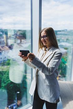 Cheerful young beautiful woman in glasses talking on a mobile phone and using a laptop with a smile while sitting at her workplace. Work in the office