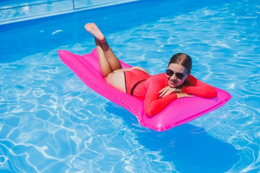 Attractive woman in sunglasses is relaxing and sunbathing on a pink mattress in the pool. Young woman in a pink swimsuit floats on an inflatable pink mattress