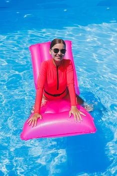 Attractive woman in sunglasses is relaxing and sunbathing on a pink mattress in the pool. Young woman in a pink swimsuit floats on an inflatable pink mattress