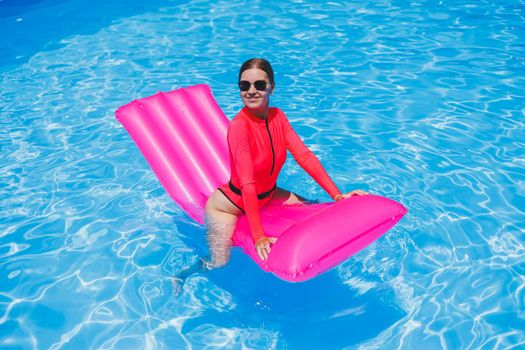 Beautiful tanned girl in a pink swimsuit and glasses by the pool. Holds a pink air mattress, sunglasses, wet tanned skin. Vacation, resort, beach. Sunny day.