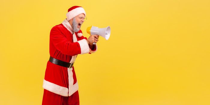 Side view of angry screaming elderly man with gray beard wearing santa claus costume holding megaphone and yelling with aggressive expression. Indoor studio shot isolated on yellow background.