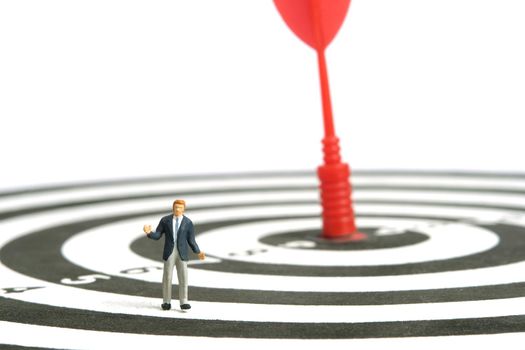 Miniature people toy figure photography. A shrugging businessman standing above dartboard with red arrow stuck in the center. Isolated on white background. Image photo