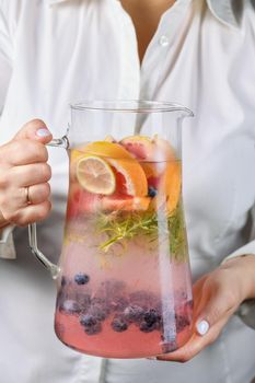 A jug with a refreshing organic soft drink with lime, blueberry, grapefruit and rosemary in female hands close-up