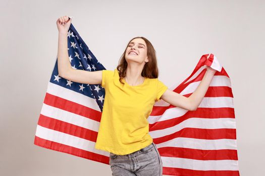 Cheerful attractive woman of young age with wavy hair, wearing yellow T-shirt holding USA flag over shoulders and keeps eyes closed and smiling happily. Indoor studio shot isolated on gray background.