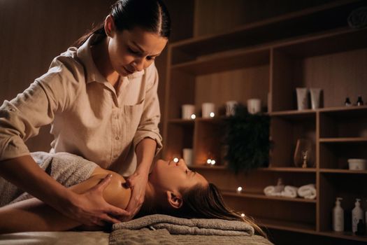 A masseuse gives a body massage to a woman in a spa center. A professional masseur massages the shoulder of a girl lying in a spa center.