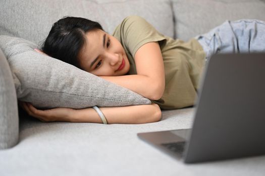 Casual young woman lying on couch and watching movie or communication through video chat on laptop.