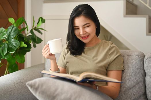 Relaxed woman drinking hot tea and reading interesting book on couch in bright living room.