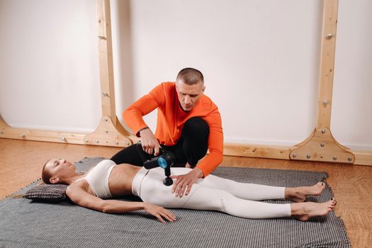 A male masseur massages a girl's body with a percussion massager to prepare for a stretching simulator.