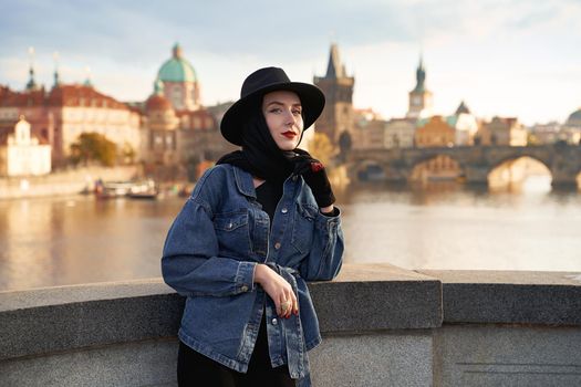 Stylish young beautiful woman earing black hat in Prague with Charles Bridge on background. Elegant retro lady fine art portrait.