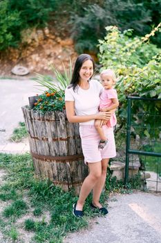 Smiling mom with a little daughter in her arms near a wooden barrel with flowers. High quality photo