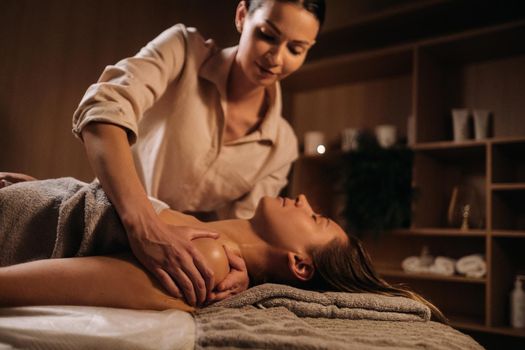 A masseuse gives a body massage to a woman in a spa center. A professional masseur massages the shoulder of a girl lying in a spa center.