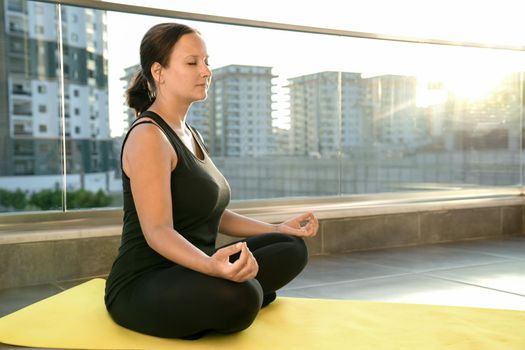 Young woman practices yoga in lotus position sitting on fitness mat on balcony of her house in rays of sun. Concept of healthy and harmonious life