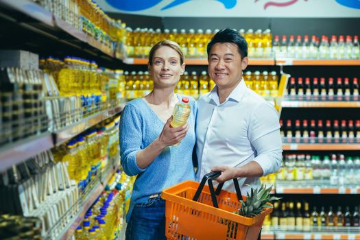 Portrait of experienced mature multiracial couple, Asian man and woman smiling and looking at camera shoppers in supermarket, choosing products, grocery department, choosing oil