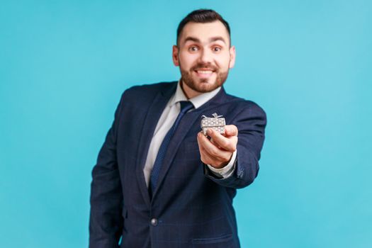 Happy smiling man wearing dark official style suit holding out little box with ring, making proposal to her girlfriend, looking at camera. Indoor studio shot isolated on blue background.