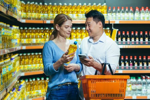 Young Asian couple using smartphone in supermarket with shopping cart choosing picking products while grocery store. browsing smart phone in food market. people make a choice oil. purchase
