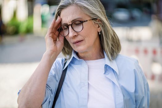 Mature grey haired woman 50s, 60s, standing outdoors holding her head suffering from migraine, headache. Suffering from summer teat feeling sick while travel mature woman standing outdoor.