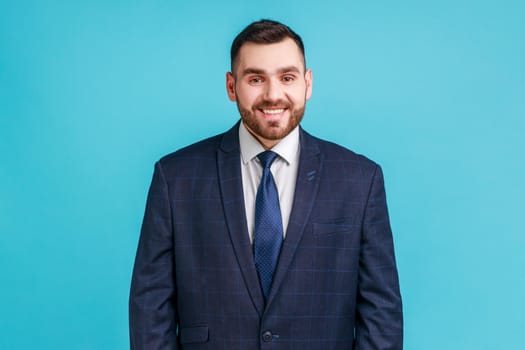 Portrait of young adult handsome businessman wearing official style suit looking at camera with toothy smile, expressing positive emotions and happiness. Indoor studio shot isolated on blue background