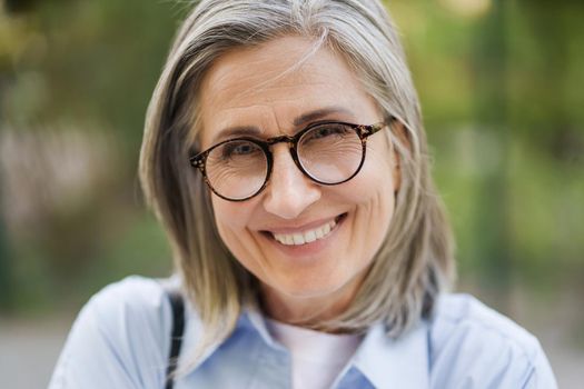 Close up portrait of charming grey hair mature woman in glasses standing outdoor enjoying free time vacation traveling around world at retirement. Mature woman with perfect skin wearing blue shirt.
