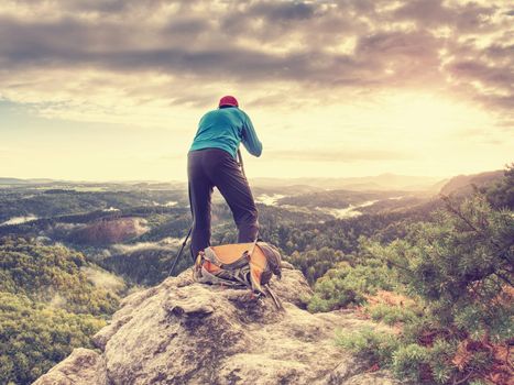 Tall photographer with baseball cap and tripod with camera in hands stand on rocky view point and watching down to deep misty valley bellow