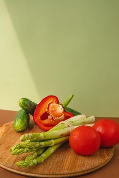 Vegetables lie on a wooden board: tomatoes, asparagus, cucumbers, red bell peppers. brown, light green background. place for text