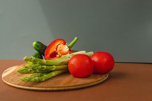 Vegetables lie on a wooden board: tomatoes, asparagus, cucumbers, red bell peppers. brown, dark gray background. place for text