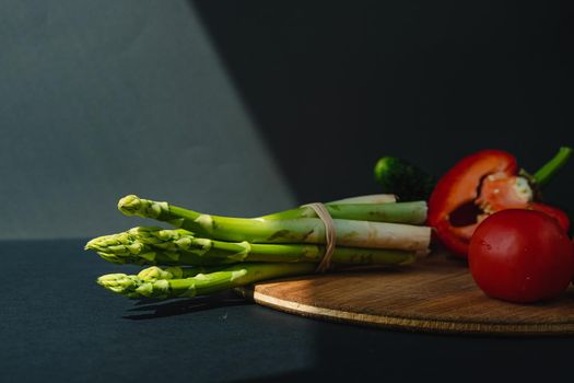 branches of fresh green asparagus, peppers and tomatoes on a wooden board, dark gray background, top view. Basic trend concept with copy space