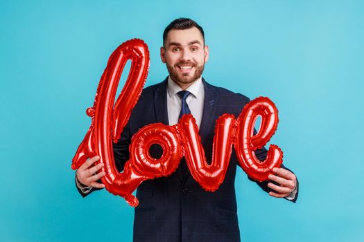 Portrait of happy smiling man wearing official style suit holding love word of foil balloons, expressing positive emotions and his feelings. Indoor studio shot isolated on blue background.